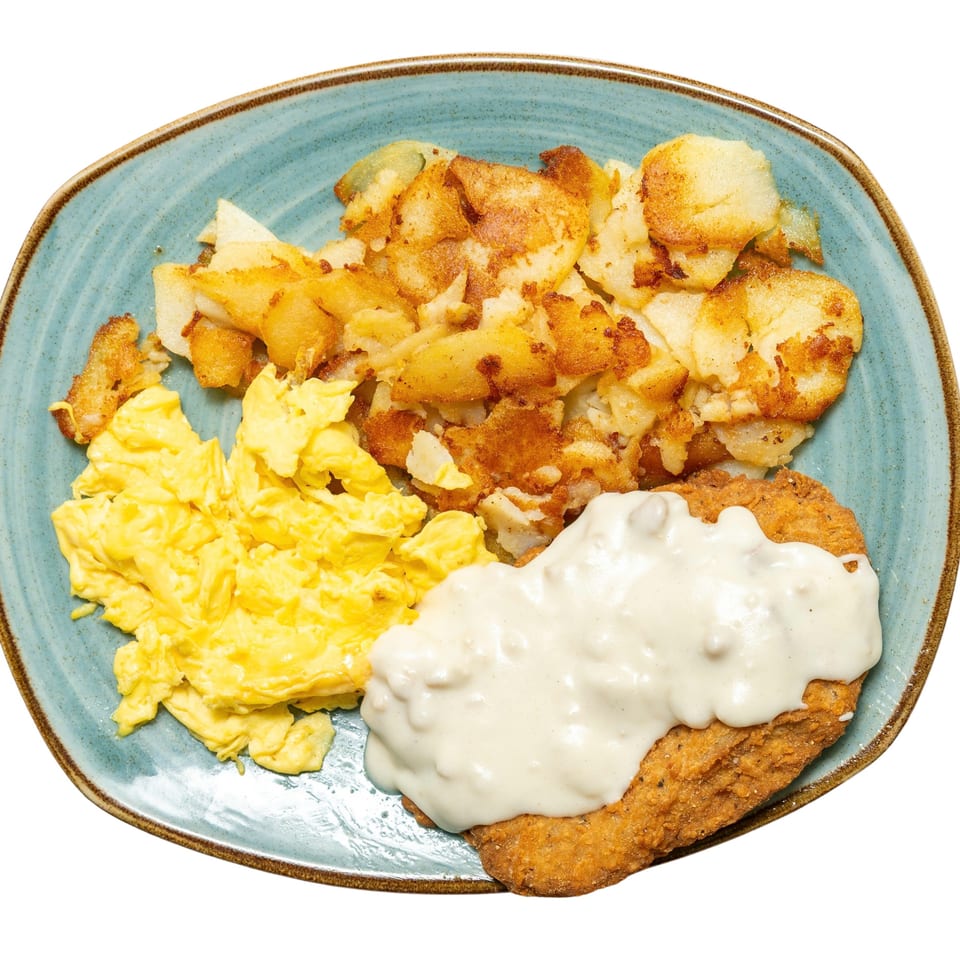 A breakfast plate with scrambled eggs, fried potatoes, and chicken fried steak with gravy.