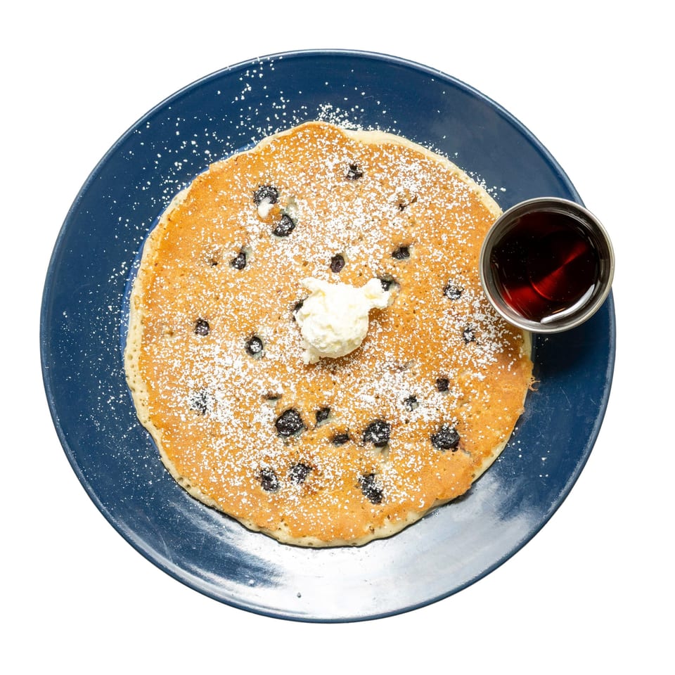 Blueberry pancake with powdered sugar, whipped cream, and a side of syrup on a blue plate.