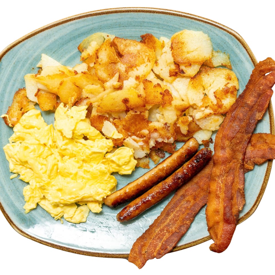 A breakfast plate with scrambled eggs, crispy home fries, two sausage links, and three strips of bacon.