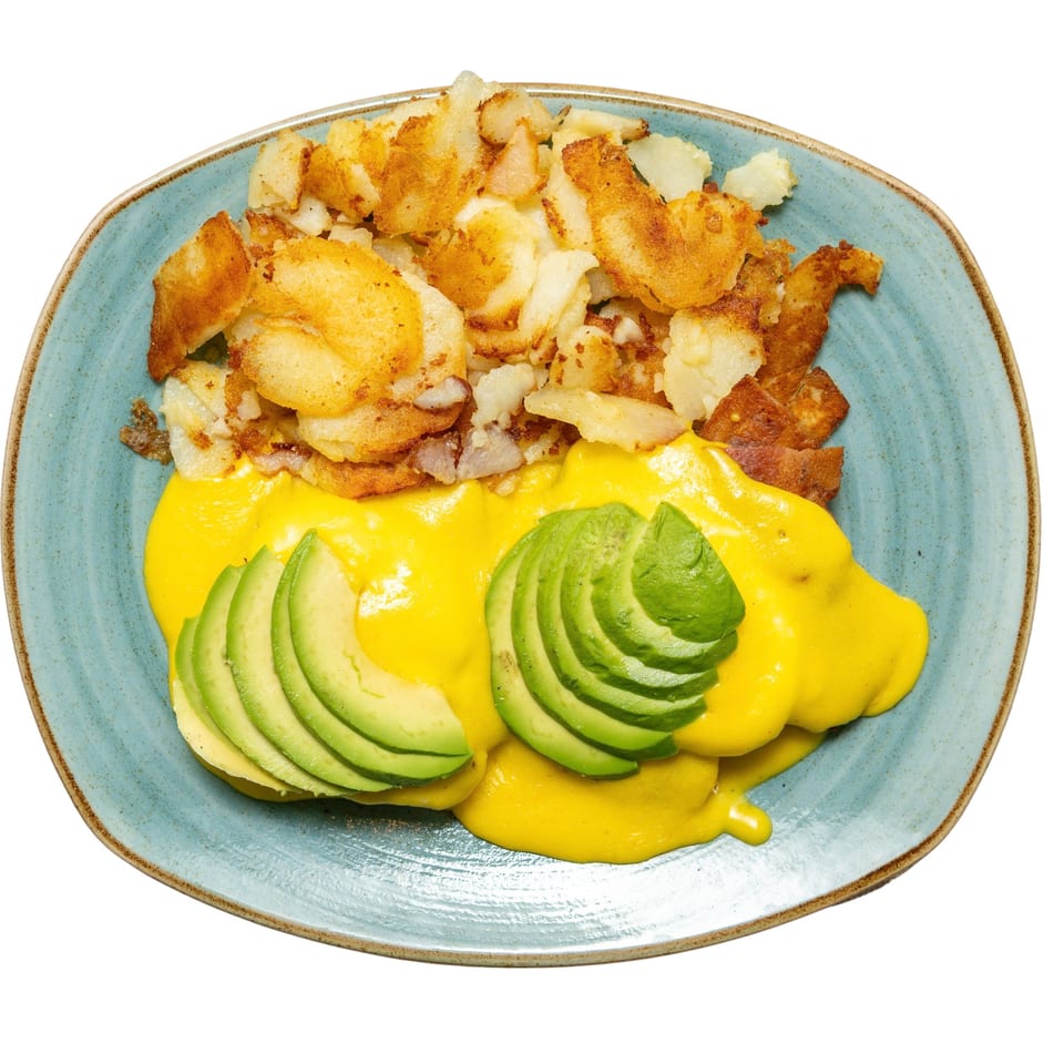 Breakfast plate with golden-brown home fries, eggs Benedict topped with hollandaise sauce and sliced avocado.